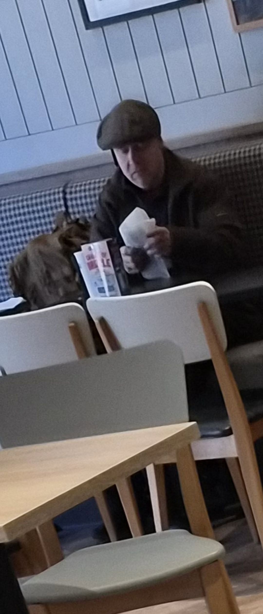 The same man seated in Greggs with a paper bag and his rucksack beside him