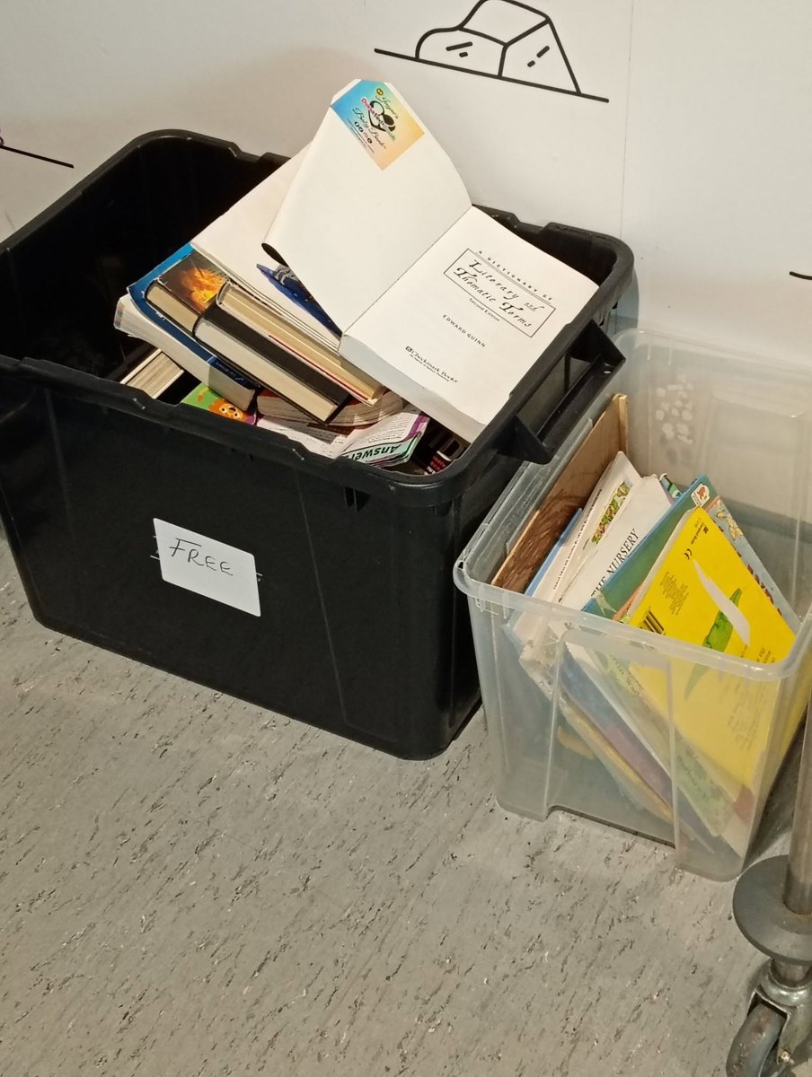 A black plastic crate labelled FREE in handwriting, filled with second-hand books with a Jayne's Baby Bank branded card on top, beside a clear tub of children's picture books on a floor