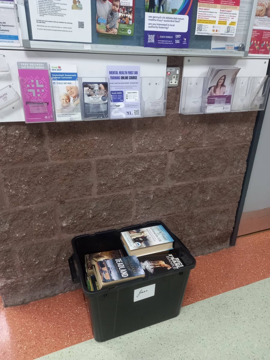 A black plastic crate of second-hand books left on the floor beneath a leaflet rack in a waiting area; the site is not confirmed